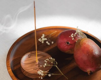 May include: A wooden incense holder with a burning incense stick, placed on a wooden tray with two ripe mangoes and small white flowers. The incense stick emits a wisp of smoke, creating a serene and natural aesthetic.