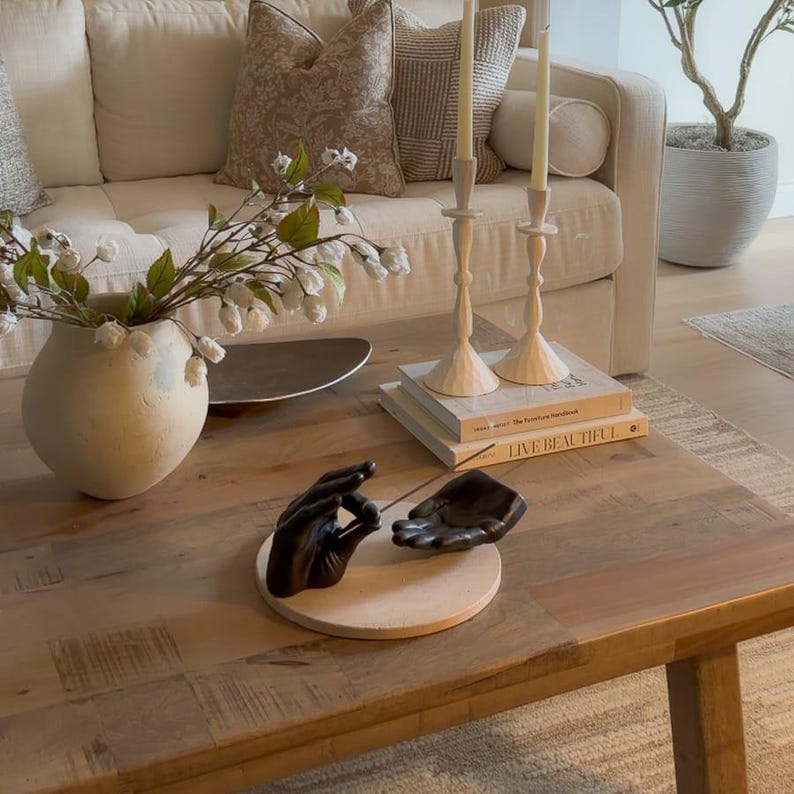 May include: A black incense holder featuring two sculpted hands resting on a circular beige base. One hand is positioned in a mudra gesture while the other is open, with a thin incense stick placed between them on a wooden coffee table.