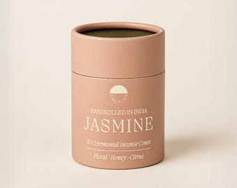 May include: A cylindrical, light pink container of jasmine incense cones. The container has a dark green interior and a white label with the word "JASMINE" in large letters. The label also states "Handrolled in India" and lists the scent notes as "Floral, Honey, Citrus."