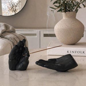 May include: A matte black incense holder set consisting of two sculpted hands. One hand is positioned upright to hold a thin wooden incense stick, while the other hand is open and flat to catch ash. The set is placed on a smooth surface.