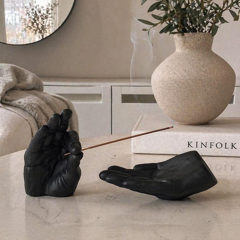 May include: A matte black incense holder set consisting of two sculpted hands. One hand is positioned upright to hold a thin wooden incense stick, while the other hand is open and flat to catch ash. The set is placed on a smooth surface.