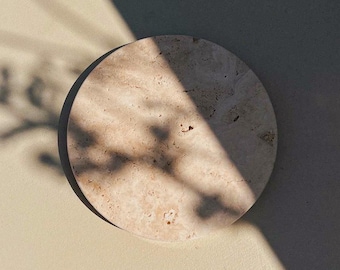 May include: A round, beige stone coaster with a textured surface. The coaster is illuminated by sunlight, casting shadows across its surface. The coaster is on a light beige surface, with shadows from a plant in the upper left corner.