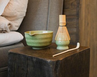 May include: A matcha tea set featuring a green ceramic bowl, a bamboo whisk, and a wooden scoop. The bowl has a spout and a textured surface. The whisk is made of bamboo and is tied with a black string. The set is on a dark wooden surface.