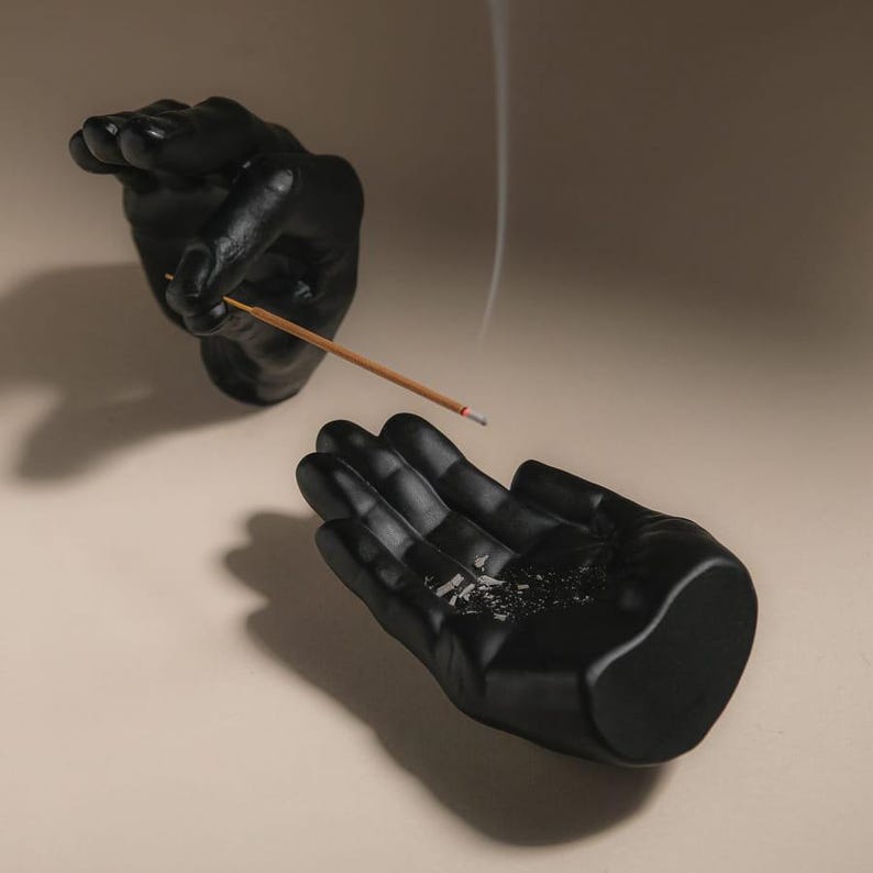 May include: A pair of matte black hand-shaped sculptures. One hand is positioned upright, holding a thin, light brown incense stick that emits a faint trail of smoke. The other hand is open, palm facing upward, serving as a tray for ash.