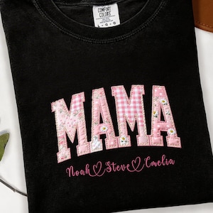 以下が含まれることがあります： ピンクの花柄とギンガムチェックのパッチワーク文字で「MAMA」と書かれた黒いTシャツ。「MAMA」の下には「Noah」、「Steve」、「Amelia」の名前が書かれています。TシャツはComfort Colors製です。