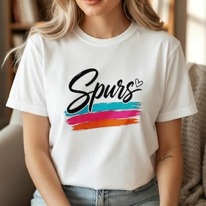 May include: A white short-sleeved t-shirt featuring the word Spurs in black cursive script with a small heart icon. Below the text are three horizontal brushstroke lines in vibrant turquoise, bright pink, and orange colors.