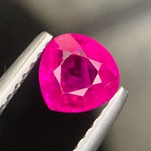 Könnte beinhalten: Ein herzförmiger, facettierter Rubin, gehalten von einer silbernen Pinzette vor dunklem Hintergrund. Der Rubin zeigt eine leuchtend rosa Farbe und interne geometrische Facetten.