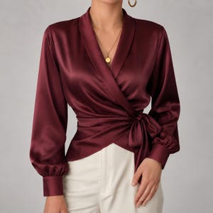 Puede incluir: Una blusa cruzada de satén color burdeos intenso con mangas largas, puños abotonados, cuello tipo chal y un lazo lateral. La tela tiene un acabado suave y reflectante. La blusa se combina con pantalones de color blanco roto.