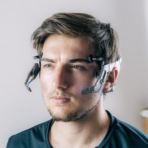 Könnte beinhalten: Eine Person trägt ein futuristisches Headset mit mehreren Sensoren und Drähten. Das Gerät ist weiß und schwarz, mit blauen Drähten. Die Person trägt ein dunkelblaues Oberteil und hat kurze braune Haare und einen Bart. Der Hintergrund ist eine schlichte weiße Wand.