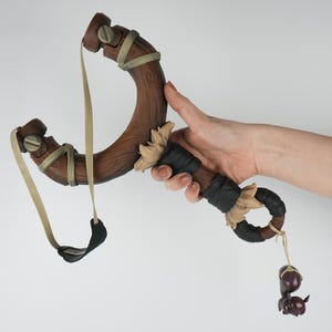 May include: A handcrafted wooden slingshot with a dark brown finish. The slingshot features a curved frame, tan elastic bands, and decorative accents. A small, dark-colored figurine hangs from the handle.