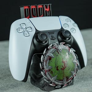 May include: A white and black PlayStation 5 controller rests on a custom stand. The stand features the word "DOOM" in red lettering and a green and red circular emblem. The controller is the primary focus, showcasing its buttons and design.