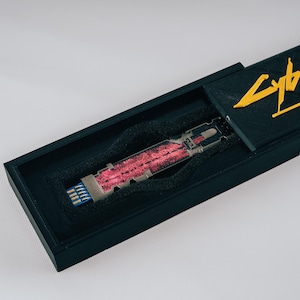 Può includere: Una scatola nera con la scritta "Cyberpunk 2077" in lettere gialle. All'interno, una chiavetta USB argentata e rossa con connettori blu, adagiata in schiuma nera. La scatola è aperta, rivelando il contenuto.