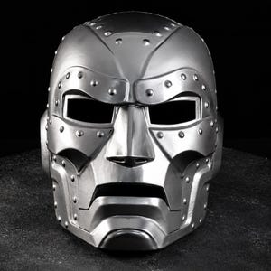 May include: A metallic silver full-face mask featuring a rigid, angular design with prominent raised rivets along the brow, cheekbones, and jawline. The mask includes rectangular eye openings and a horizontal mouth slit, finished with a smooth sheen.
