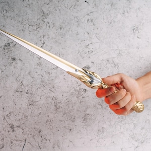 May include: A decorative dagger with a gold-colored handle and a silver blade. The handle features ornate detailing, and the blade is sharp and pointed. The dagger is held against a gray background.