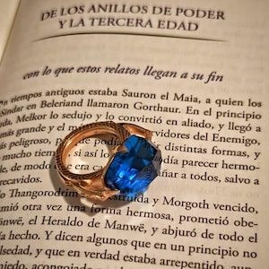 May include: A gold-toned metal ring featuring an intricate, textured band and a large, faceted, rectangular blue gemstone. The ring is resting on an open book page with Spanish text titled DE LOS ANILLOS DE PODER Y LA TERCERA EDAD.