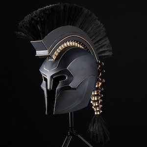 May include: A matte black Spartan-style helmet featuring a prominent black crest made of stiff bristles. The helmet is accented with gold-coloured metallic details along the ridge and a decorative gold-coloured spine-like structure at the back.