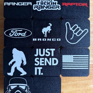 May include: A collection of black square coasters featuring various white and red graphics and text. Designs include the Ford logo, Ranger, Raptor, Bronco, a hand gesture, the American flag, a Bigfoot silhouette, a Stormtrooper helmet, and the phrase "JUST SEND IT."