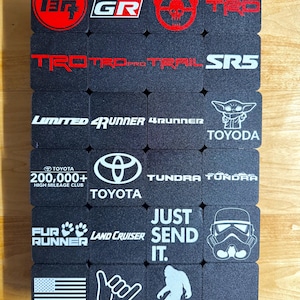 May include: A collection of black square coasters with various white and red graphics and text. Logos include "TRD", "GR", "Toyota Racing Development", "TRD Pro Trail", "4Runner", "Toyota", "Just Send It", and a Stormtrooper.