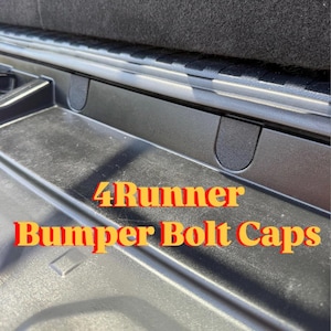 Può includere: Tappi per bulloni del paraurti in plastica nera per un 4Runner. L'immagine mostra un primo piano dei tappi installati nell'area del paraurti di un veicolo. Il testo "4Runner Bumper Bolt Caps" è visualizzato in arancione e rosso.