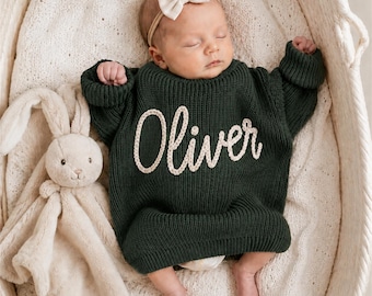 May include: A dark forest green long-sleeve knit sweater featuring the name Oliver embroidered in cream-colored yarn across the chest. The sweater is worn by a sleeping infant who is also wearing a cream-colored bow headband next to a plush toy.
