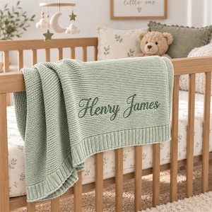 May include: A sage green knitted baby blanket featuring a ribbed border and the name Henry James embroidered in a dark green cursive script. The blanket is draped over the wooden railing of a crib, showcasing its textured garter stitch pattern.