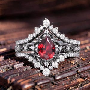 May include: A three-piece ring set featuring a pear-shaped, deep red gemstone at the center. The ring is surrounded by smaller, clear gemstones and intricate metalwork. The bands are silver-toned, creating a striking contrast with the red gemstone.