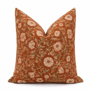 May include: A square throw pillow featuring a burnt orange base color with a repeating pattern of light beige daisy-like flowers and muted green foliage. The fabric has a visible woven texture throughout the entire surface of the pillow cover.