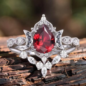 May include: A silver engagement ring set with a large, pear-shaped, deep red gemstone. The ring is surrounded by smaller white and black gemstones, with intricate detailing on the band. The ring is set on a wooden surface.