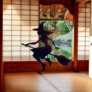 May include: A dark silhouette of a figure wearing a pointed hat and high-heeled shoes, riding a broomstick. The figure is positioned against a bright shoji screen window that overlooks a pond and trees in a Japanese-style room with tatami mats.