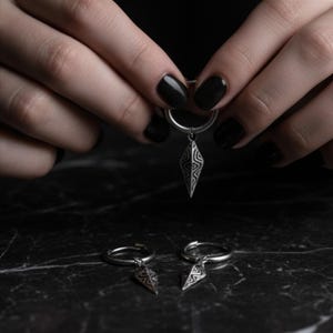 May include: Silver-toned earrings with a geometric pendant. The earrings are held by a person with black nail polish. Two additional earrings are visible on a dark surface, showcasing the design and style of the jewelry.