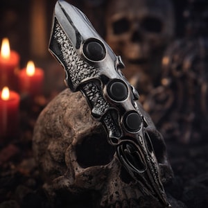 Puede incluir: Anillo de armadura para dedo plateado con tres acentos circulares negros. El anillo tiene un diseño intrincado y texturizado y se coloca sobre un fondo borroso con una calavera y velas encendidas, creando una estética gótica.