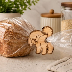Op de afbeelding: Een lichtbruine houten broodclip in de vorm van een poedel met een krullende staart. De clip is bevestigd aan een doorzichtige plastic zak met een bruin brood. Het poedelontwerp is voorzien van gegraveerde contouren van het gezicht en lichaam.