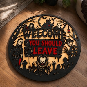 May include: A circular wooden wall sign featuring black silhouettes of horror movie characters and a haunted house. The text reads WELCOME in black and YOU SHOULD LEAVE in red. The design includes figures like a clown, a masked slasher, and a zombie.