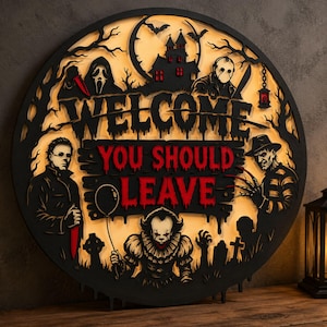 May include: A circular black wooden wall sign with a backlit yellow background featuring horror movie characters. The text reads WELCOME YOU SHOULD LEAVE in black and red. It depicts figures including a clown, a masked slasher, and a haunted house.