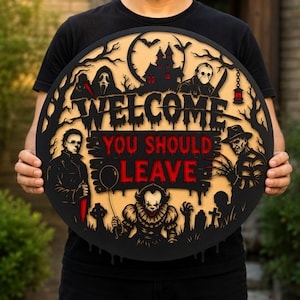 May include: A circular black and beige wooden sign featuring the text WELCOME YOU SHOULD LEAVE in red and black. The design includes layered silhouettes of horror movie characters, a haunted house, bats, trees, and tombstones in a dark theme.