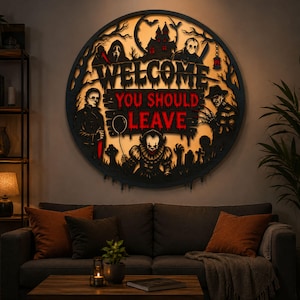 May include: A circular black wall sign featuring a backlit horror-themed design with the text "WELCOME YOU SHOULD LEAVE" in red and black. The artwork includes silhouettes of iconic horror movie characters, a haunted house, and a graveyard.