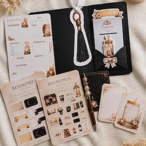 Könnte beinhalten: Ein Schreibwaren-Set bestehend aus einem Bookoria Lesetagebuch, einer schwarzen Mappe mit Quasten-Lesezeichen, Stickerbögen mit Buch- und Kaffeemotiven, einem Stift mit Perlen sowie kleinen Notizkarten auf einem strukturierten Stoffuntergrund.
