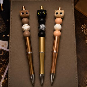 May include: Three pens featuring cat-shaped toppers and decorative beads. The left pen is bronze with brown and white beads, the center pen is gold with black and marbled beads, and the right pen is rose gold with peach and white beads.