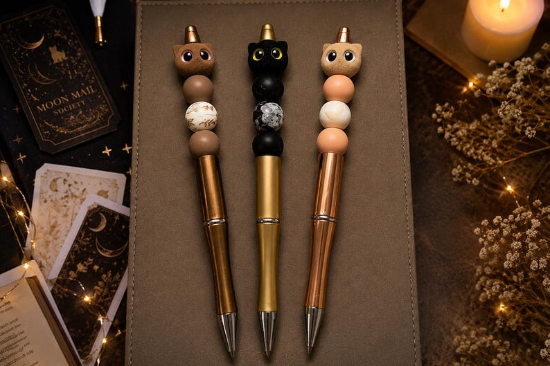 May include: Three pens featuring cat-shaped toppers and decorative beads. The left pen is bronze with brown and white beads, the center pen is gold with black and marbled beads, and the right pen is rose gold with peach and white beads.
