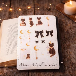 May include: A rectangular sticker sheet titled Moon Mail Society featuring illustrations of black cats, crescent moons, keys, and black bows. The sheet includes various sizes of these motifs, with some cats sitting, some reading, and others adorned with bows.