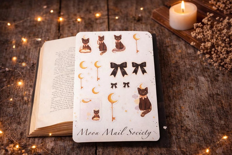 May include: A rectangular sticker sheet titled Moon Mail Society featuring illustrations of black cats, crescent moons, keys, and black bows. The sheet includes various sizes of these motifs, with some cats sitting, some reading, and others adorned with bows.