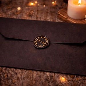 May include: A rectangular black envelope featuring a circular wax seal at the center of the flap. The wax seal is black with gold-colored detailing that depicts celestial motifs including a crescent moon and stars, creating a distinct contrast.