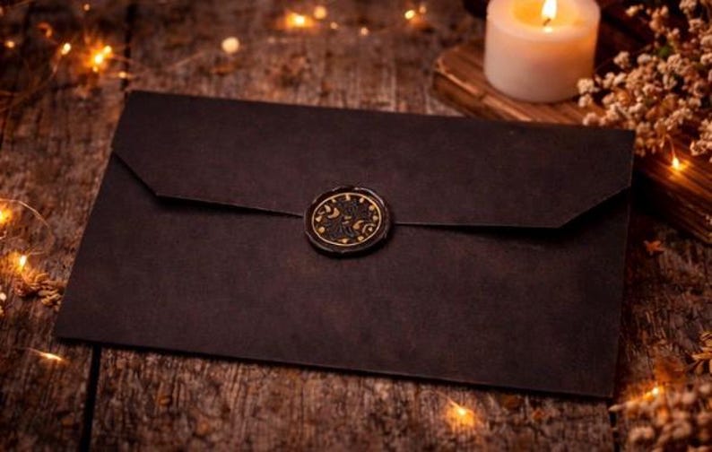 May include: A rectangular black envelope featuring a circular wax seal at the center of the flap. The wax seal is black with gold-colored detailing that depicts celestial motifs including a crescent moon and stars, creating a distinct contrast.