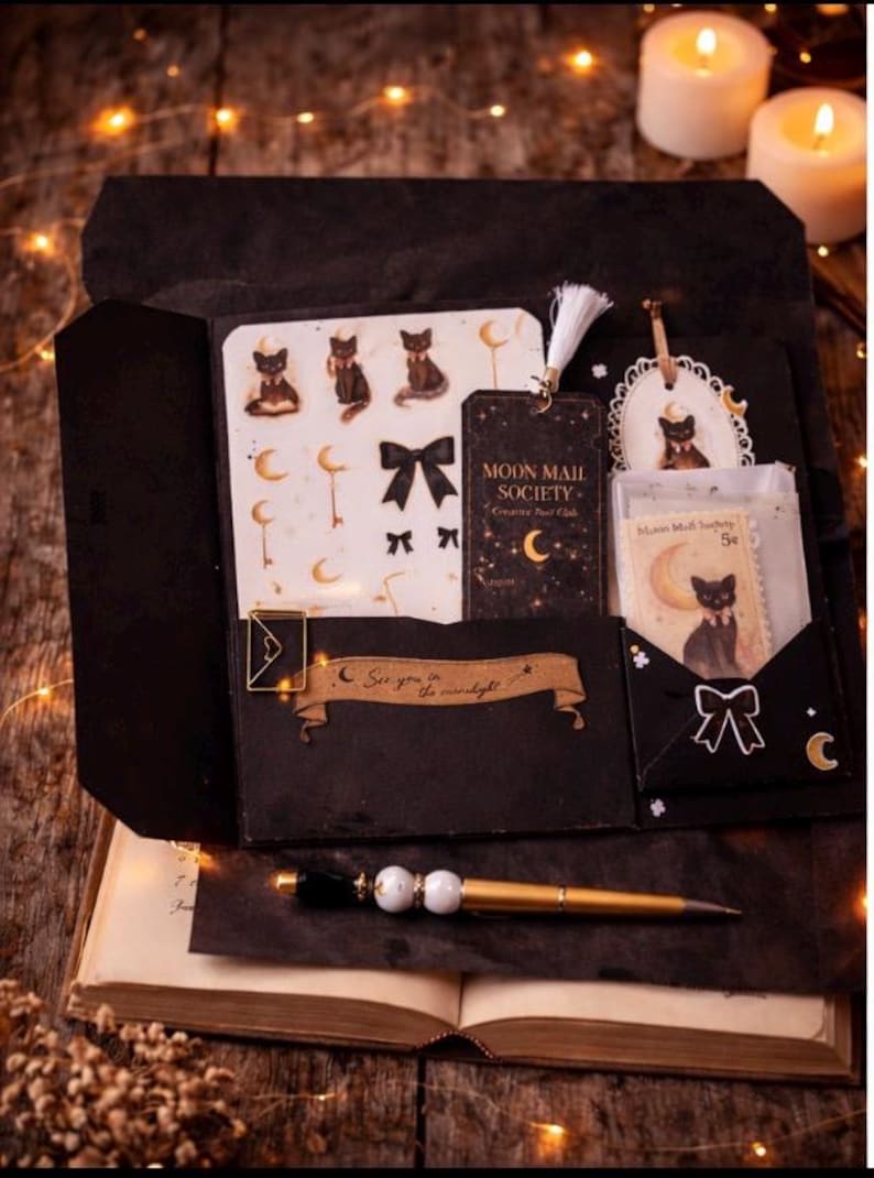 May include: A black stationery folder containing a sheet of cat and moon stickers, a Moon Mail Society tag with a white tassel, a small envelope with a cat illustration, and a gold-toned pen. The folder features a banner with the text See you in the moonlight.