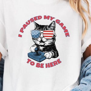 May include: White t-shirt featuring a black and white cat wearing sunglasses with American flag designs. The cat is holding a blue game controller. Text around the cat reads "I PAUSED MY GAME TO BE HERE" in red.