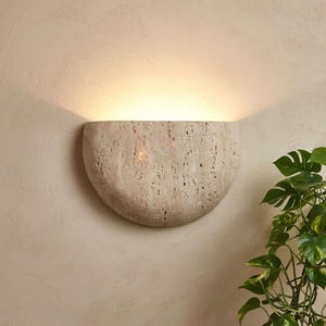 May include: A beige, semi-circular wall sconce made of textured stone. The sconce emits a warm, diffused light, casting a soft glow on the wall. A green plant with large leaves is visible in the lower right corner.