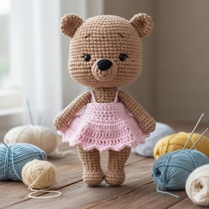Peut inclure: Poupée ours au crochet faite à la main en fil beige, vêtue d'une robe rose clair. L'ours a un nez et des yeux noirs. La poupée est posée sur une surface en bois avec des pelotes de laine en arrière-plan.