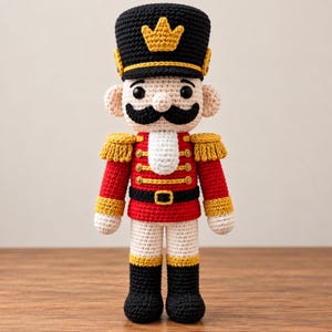 May include: A crocheted nutcracker doll featuring a black hat with a yellow crown, a red jacket with gold trim and buttons, a white chest piece, black boots, and a prominent black moustache. The figure has light skin and black bead eyes.