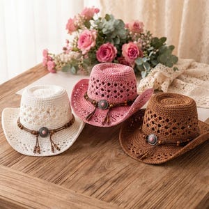Könnte beinhalten: Drei Stroh-Cowboyhüte in Weiß, Pink und Braun. Jeder Hut hat eine gehäkelte Krone mit Rautenmuster, eine geschwungene Krempe und ein dekoratives Band aus brauner Kordel, Holzperlen und einem mittigen, silberfarbenen Metall-Concho.