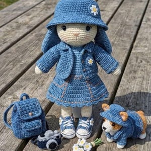 Puede incluir: Una muñeca de ganchillo con un conjunto estilo vaquero azul que incluye sombrero de pescador, chaqueta y falda a juego, decorado con margaritas blancas. La muñeca lleva zapatillas azules y blancas. Junto a ella hay una mochila, una cámara y un corgi.